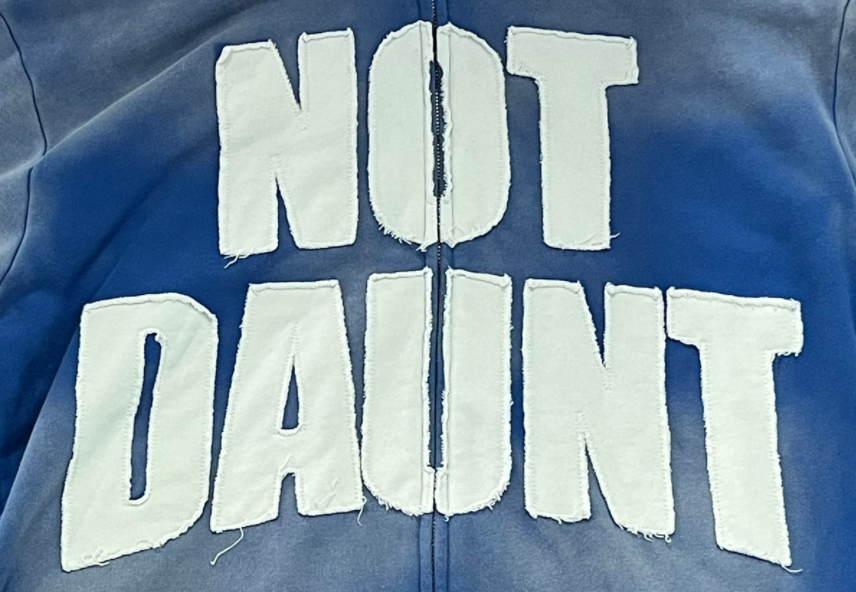 Applique embroidery sample with white frayed letter patches spelling NOT DAUNT on gradient blue zip-up jacket