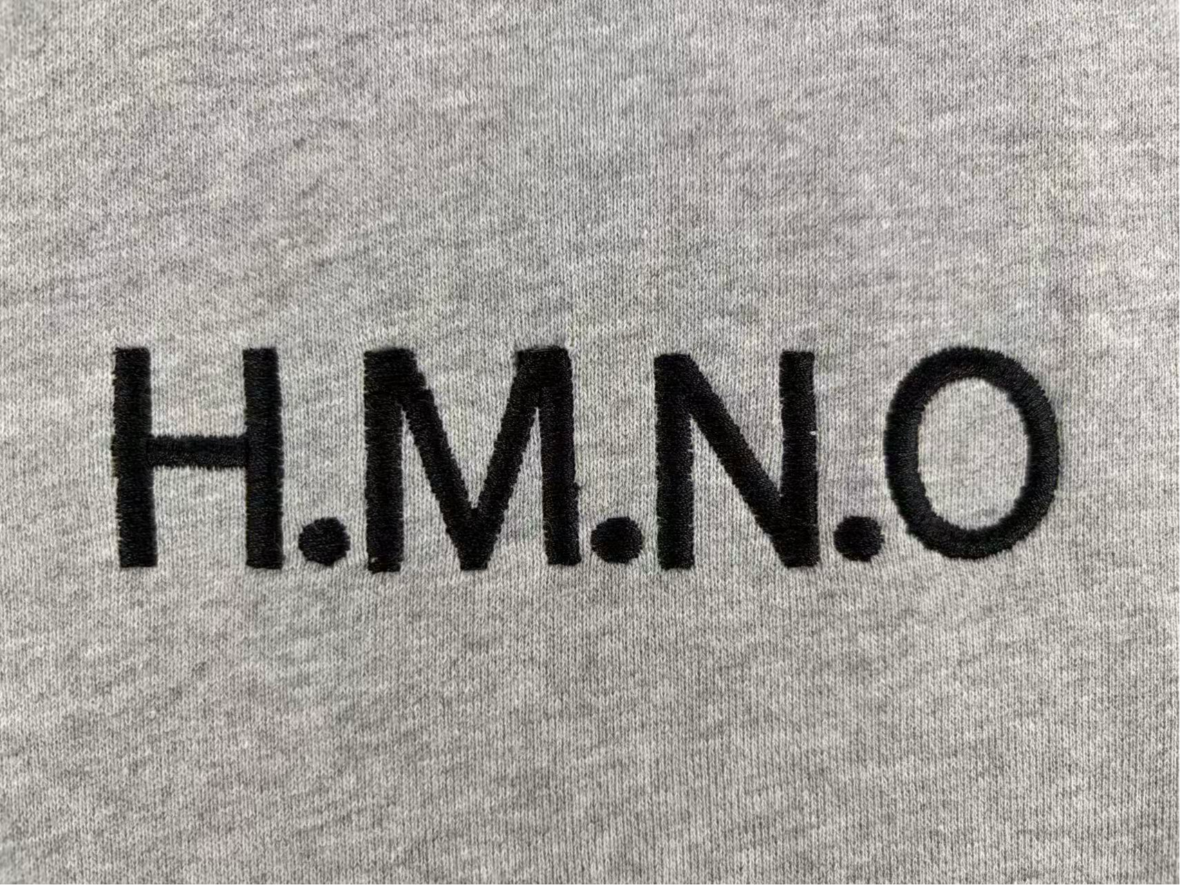 Close-up of black embroidered text H.M.N.O on grey heather fabric, flat embroidery sample detail